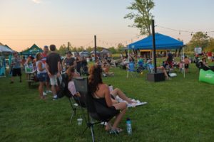 A group of park visitors sit in foldable lawn chairs on the lawn, surrounded by tents and other visitors enjoying a fun evening event in the park.